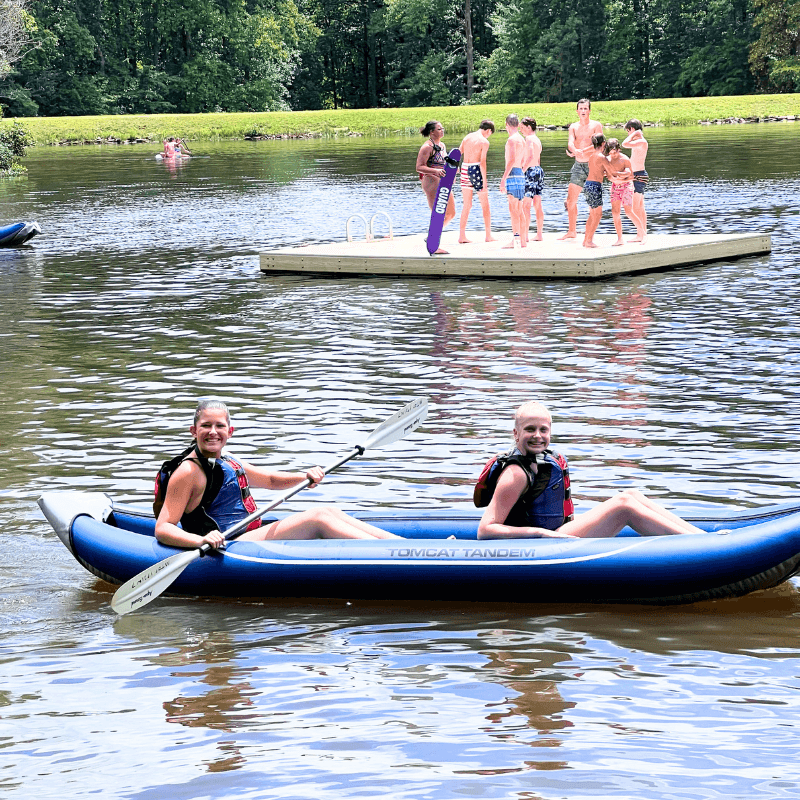 two girls kayaking on the lake, while a group of kids behind them stand on a floating dock at signature sports camp