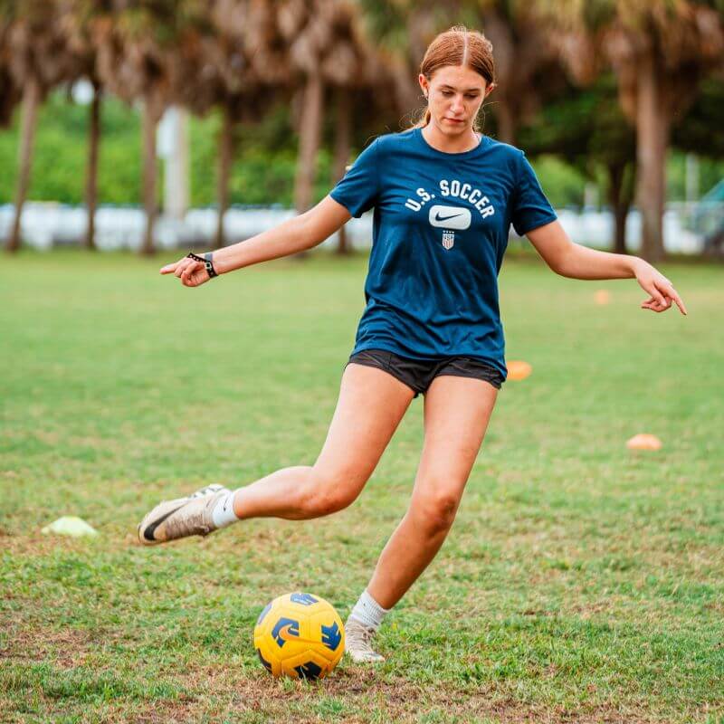 Girl wearing a blue US Soccer shirt is dribbling a yellow soccer ball at sports camp