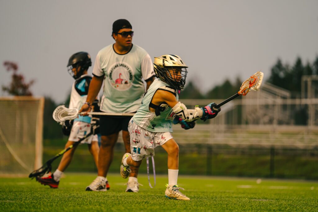 Lacrosse players in light uniforms compete on a green field; a goalie in a helmet guards the goal in the foreground.
