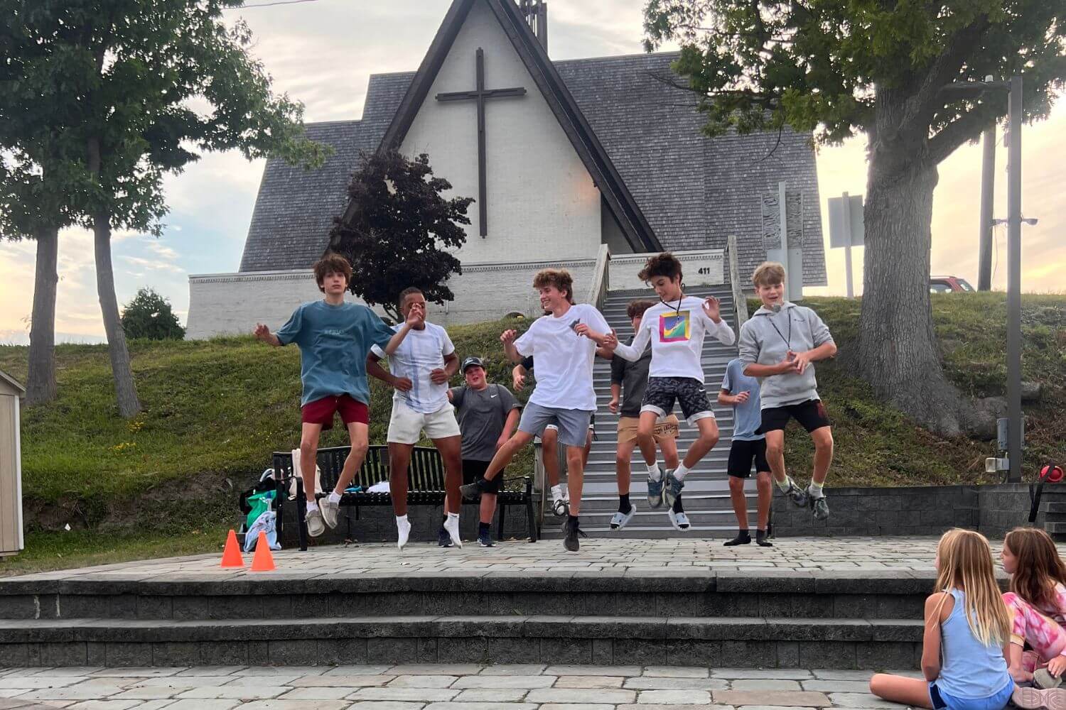 five boys jumping in the air on an outdoor stage while friends look on