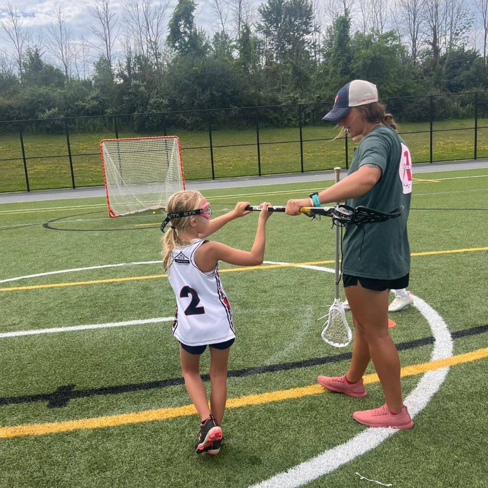 A female professional lacrosse player is coaching a young lacrosse athlete during summer camp