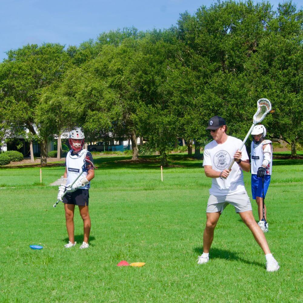 A lacrosse coach is providing a skill demonstration to a lacrosse player on an outdoor field