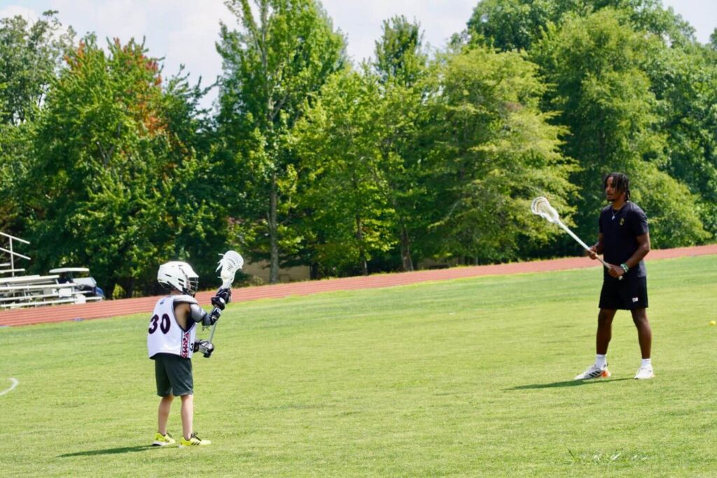 A young lacrosse player is getting one one-on-one lacrosse fundamentals training with a professional coach on an outdoor field.