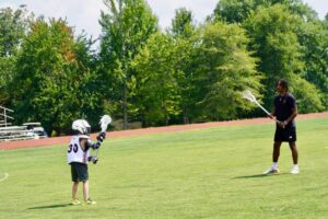 A young lacrosse player is getting one one-on-one lacrosse fundamentals training with a professional coach on an outdoor field.