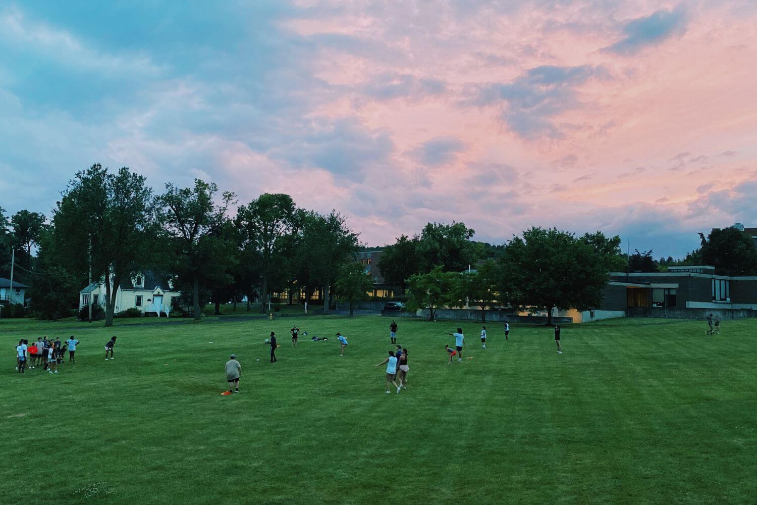 Group of summer campers are on a large field at sunset playing a game of kickball