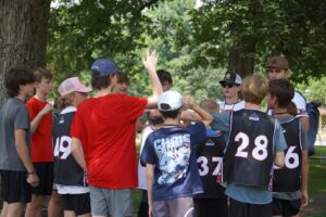 A group of boys at summer camp huddled together under a tree, one boy is raising his hand