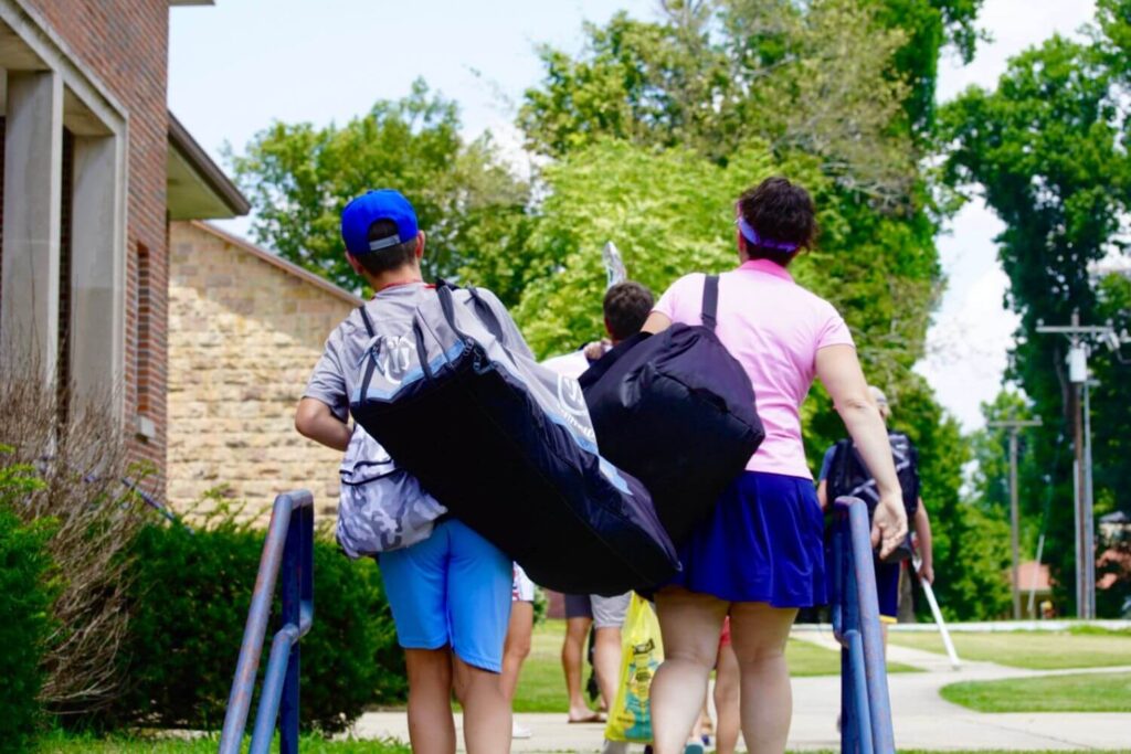 A male and female lacrosse player are walking into overnight sports camp, carrying their duffel bags and lacrosse equipment