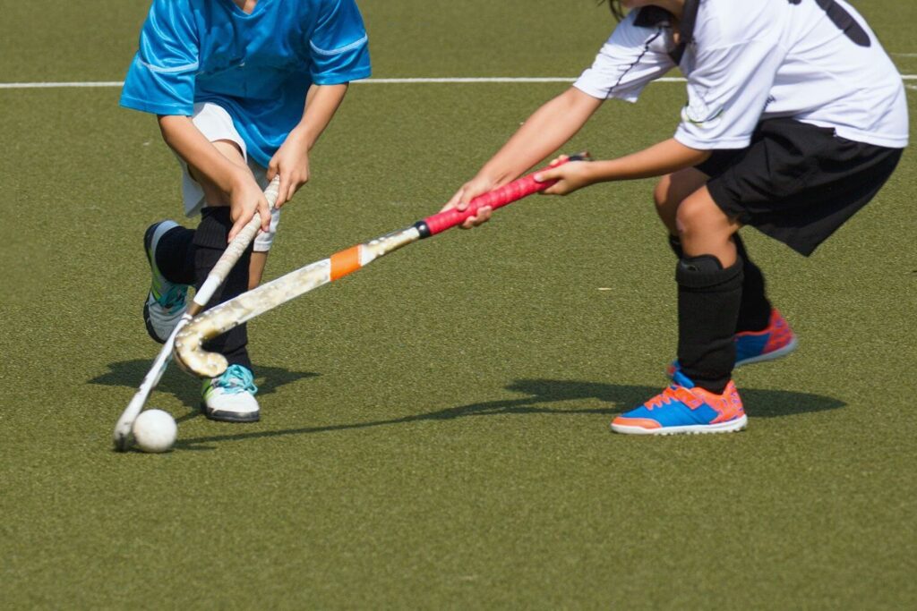 Two child field hockey players competing, attacker in blue jersey and defender in white jersey