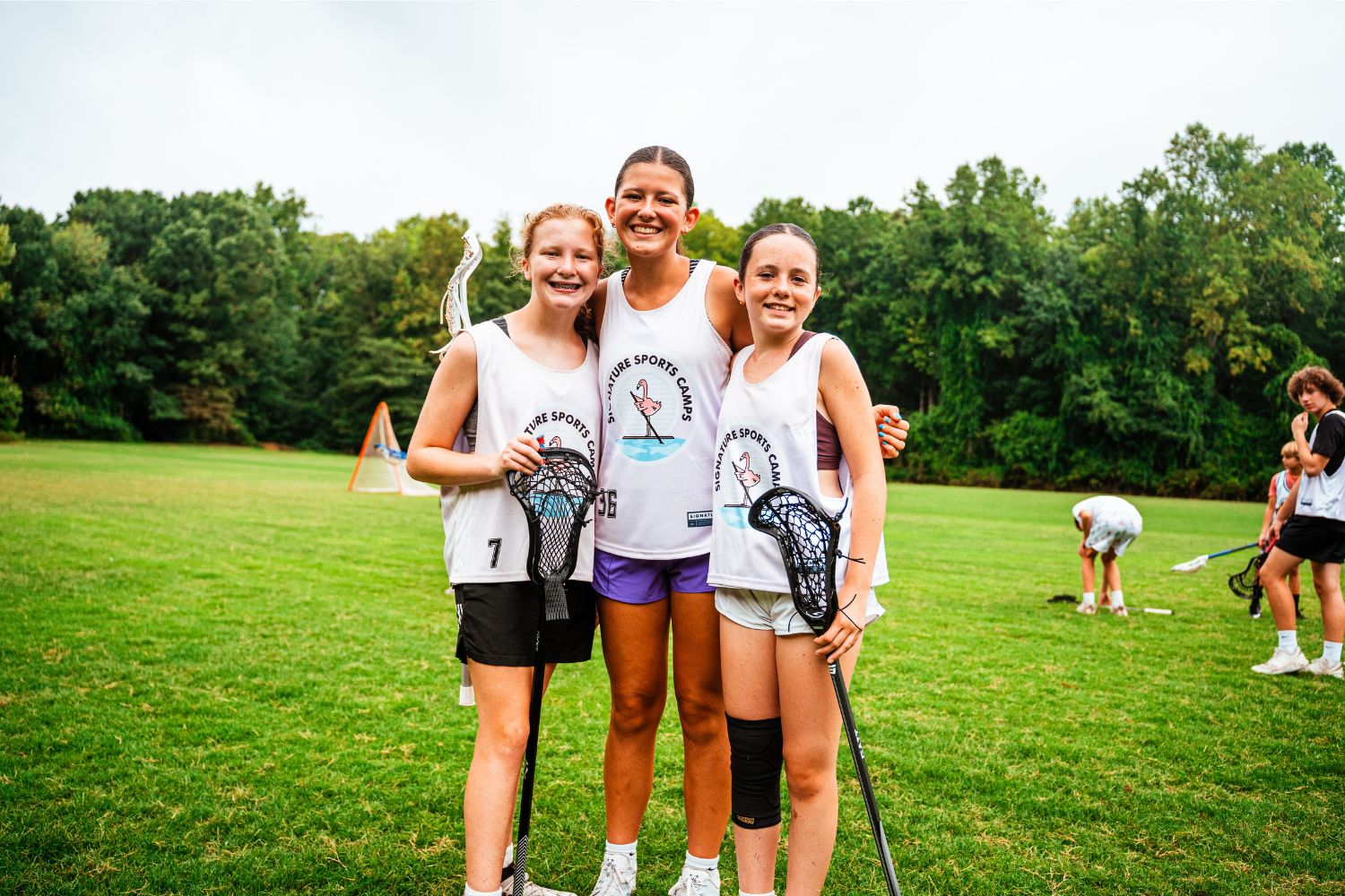 Three girls wearing Signature Sports Camp uniforms holding lacrosse sticks