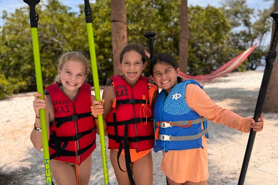 Three girls in life jackets holding paddleboard oars smile for a photo on the beach