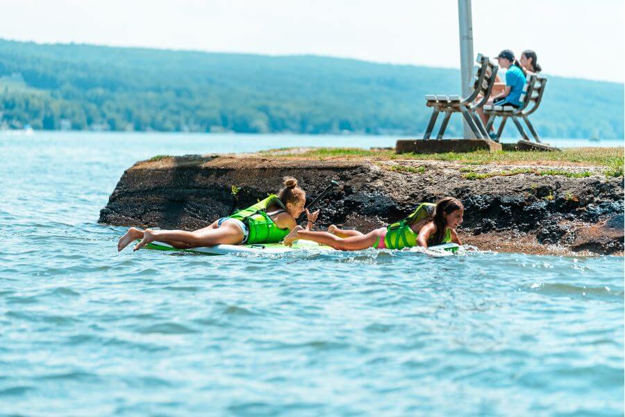 Girls wearing lifevests laying face down on paddleboards on the lake