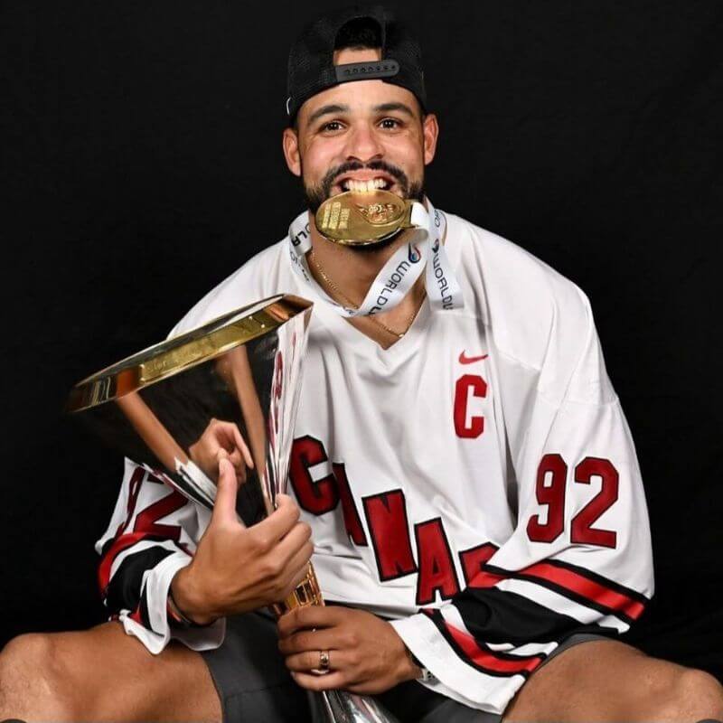 Pro lacrosse player Dhane Smith wearing Canada jersey, holding a trophy and a gold medal in his mouth after winning lacrosse championship