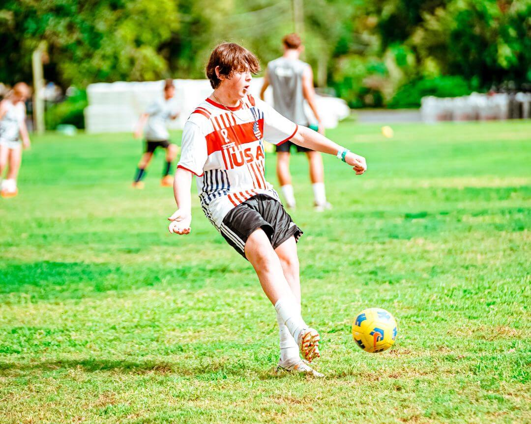 Boy in a team USA soccer jersey dribbles a yellow soccer ball at sports camp