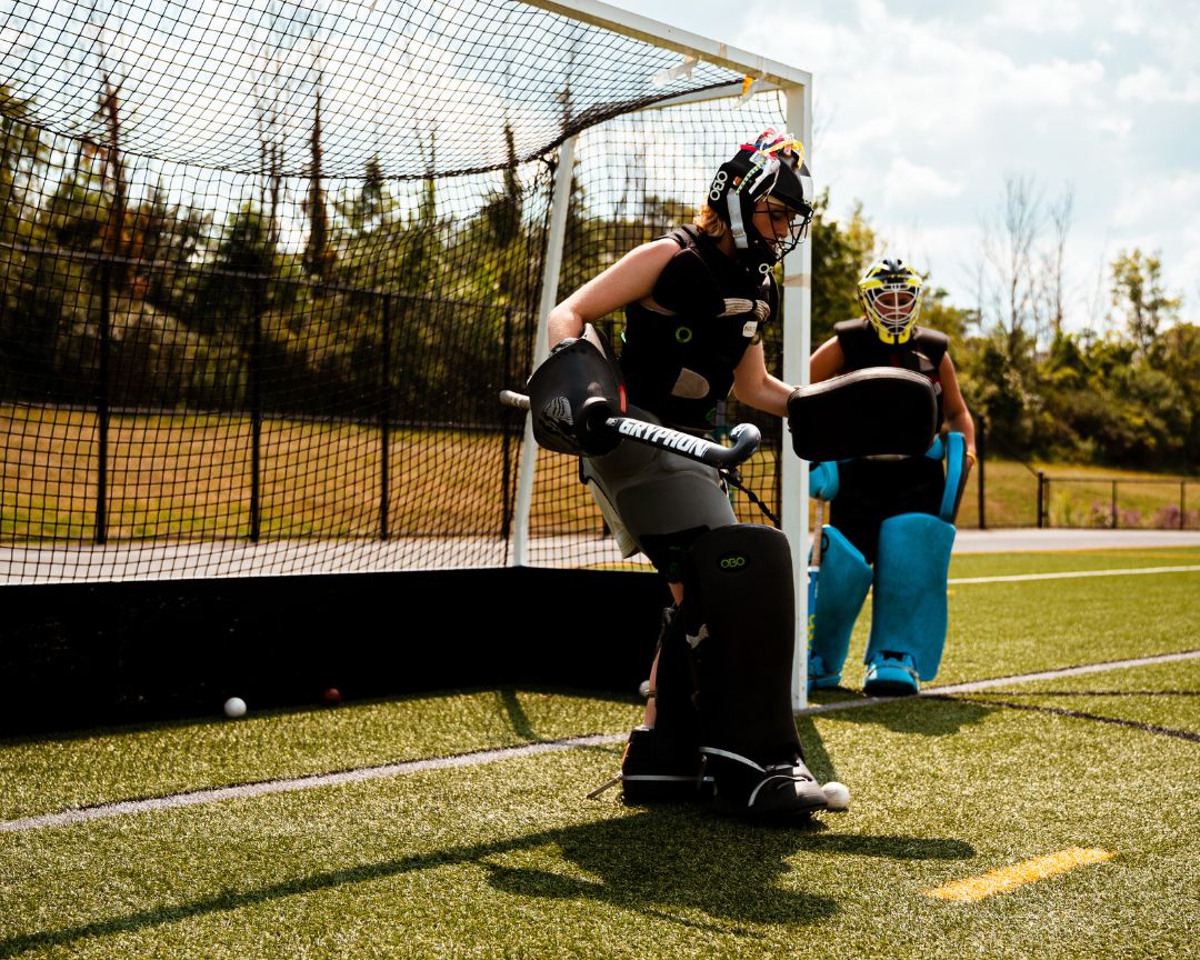 Field hockey goalie padded up standing in front of a goal box during camp practice