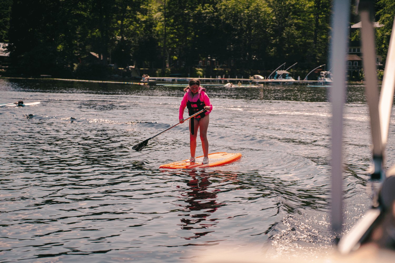 girl in a pink life jacket stand up paddleboarding on a lake at sports camp