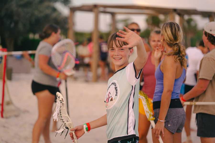 boy at overnight sports camp waves to the camera while holding his lacrosse stick