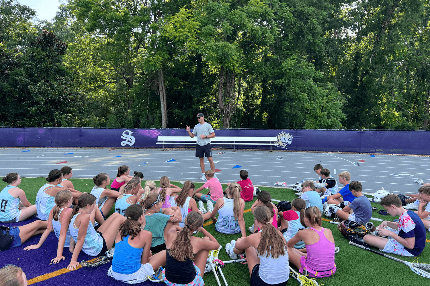 Lacrosse players sit on the field as a lacrosse pro speaks to them at overnight sports camp